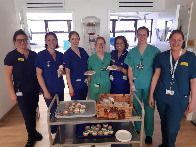 Seven smiling healthcare professionals in scrubs stand around a trolley of cupcakes in a hospital room.
