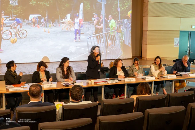 Sept personnes sont assises à une table de conférence dans un amphithéâtre, avec la secrétaire générale, Nicole Charbonnier, debout au centre tenant un micro. Plusieurs papiers et gobelets sont posés sur la table devant elles. Un grand écran en arrière-plan affiche une photo d’activités sportives de rue, où des enfants jouent au basket et au vélo. Quelques personnes du public sont visibles de dos, assises sur des fauteuils de conférence. L’ambiance est professionnelle et studieuse.