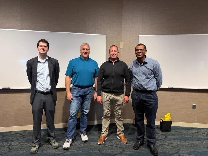 Dr. Andrew Swartz, Howard Herrild, Brett Stanton and Baron Colbert stand side by side in a conference room in front of two whiteboards. They are dressed in business casual attire and smiling at the camera. The group is positioned against a neutral-colored wall, with a small gift bag visible on the floor to the right.