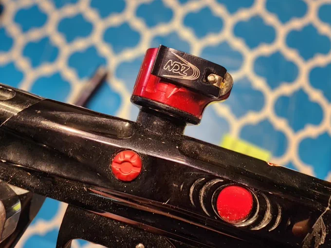 Close-up of an early 2000s NDZ paintball part in red anodized aluminum attached to a black paintball marker, with visible wear and machining details.