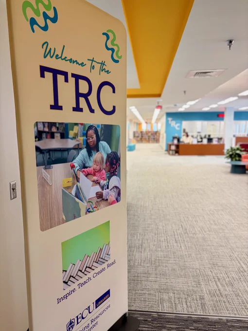 A sign welcoming visitors to the TRC, displaying the slogan "Inspire. Teach. Create. Read." with a photo of an individual assisting children with books. The interior further shows a hallway leading to the Teaching Resources Center entrance. 