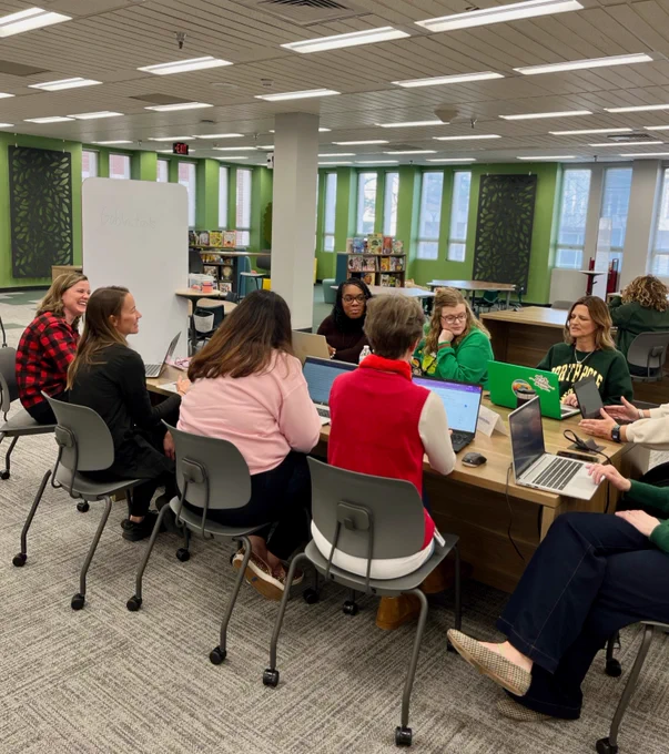 A group of people sitting around a table with laptops in a library setting. There is a whiteboard with writing in the background and bookshelves along the walls.