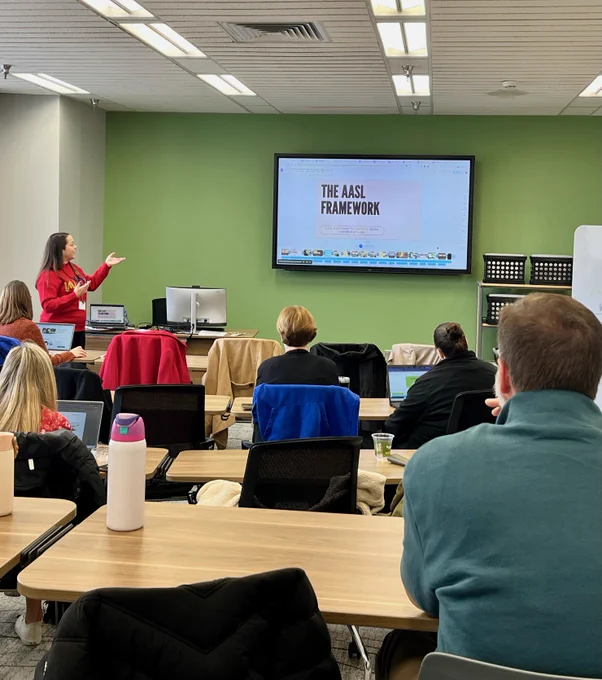 A person stands at the front of a classroom, giving a presentation on "The AASL Framework" displayed on a screen. Several people are seated at tables, engaging with the presentation. The room has a bright and modern setting.