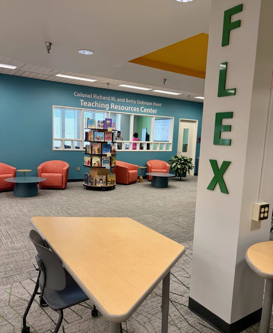 Library interior with a sign reading "Colonel Richard M. and Betty Debnam Hunt Teaching Resources Center" on a blue wall. Comfortable chairs are arranged around a small shelf filled with books. A pillar at the foreground has green letters spelling "FLEX.