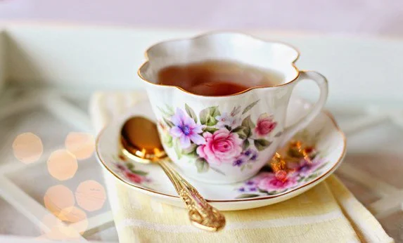 A floral teacup and saucer with a golden spoon, filled with tea, on a yellow cloth with bokeh lights.