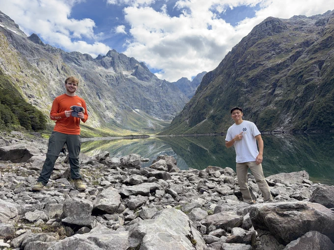 Two people stand on rocky terrain beside a serene mountain lake, surrounded by steep, green hills under a partly cloudy sky.