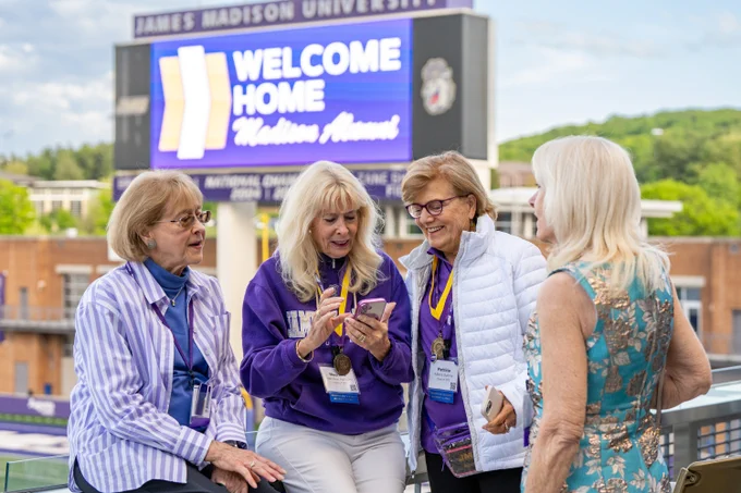 Three alums look at something on a cellphone. Behind them on the Bridgeforth Stadium score board has a graphic that reads “Welcome Home Madison Alumni”.