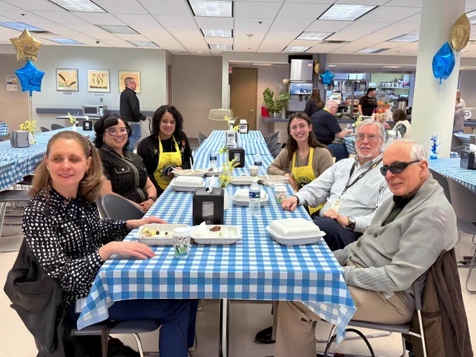 A group of people sit around a table covered with a blue and white checkered tablecloth in the cafeteria.  The room is decorated with blue and gold star balloons.