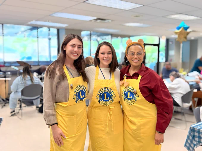 Grace, Lindsay, and Angela K. stand together wearing yellow aprons with the Lions Club International logo. Balloons are visible in the background.