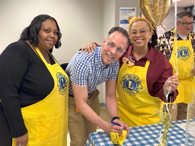 A group of people standing at a table covered with a blue and white checkered cloth. They are wearing yellow aprons with the "Lions Clubs International" logo. One person is signing a ticket while others smile beside a gold balloon.