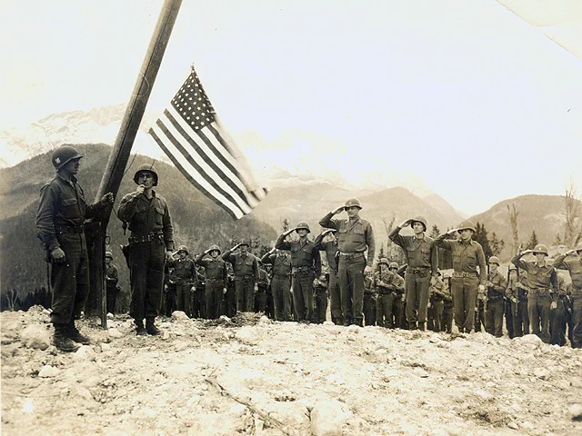 Historic photo of U.S. Soldiers raising the American flag near Bad Reichenhall in 1945 as troops stand in formation and salute during the liberation.