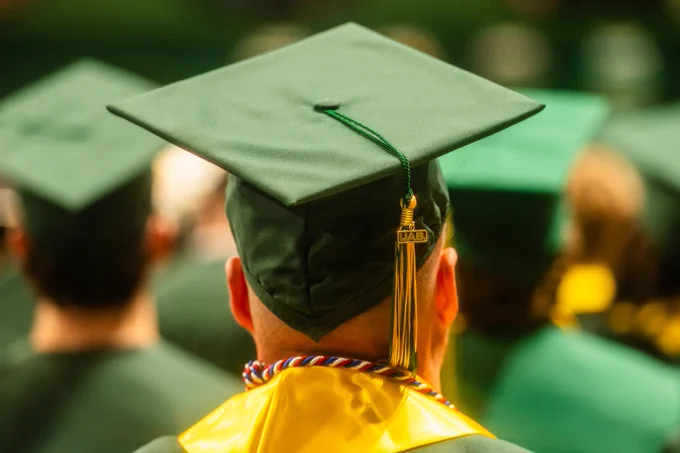 A person wearing a green graduation cap and gown is seen from behind. The cap is adorned with a green and gold tassel, and the person has a multicolored cord around their neck. A crowd of graduates is blurred in the background.