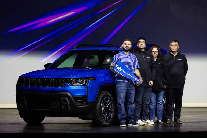 Five people stand in front of a blue car, holding a sign that reads \"University at Buffalo\". They're on a stage with purple lighting behind them.