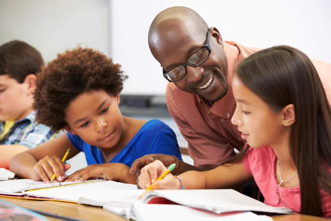 A teacher helps two young students with their work at a desk in a classroom.