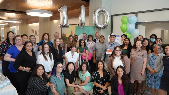A group of people posing indoors next to large balloons forming the number "100." Some hold certificates, and there are balloons in various colors behind them. 