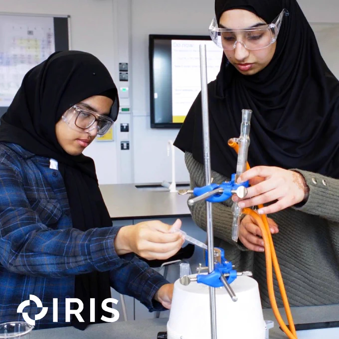Two students are conducting a lab experiment in a classroom, using a stand, test tubes and clamps. The IRIS logo is in the bottom left corner