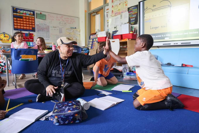 A smiling teacher sitting on a carpet in a classroom giving a high-five to a smiling young student. They are in a classroom surrounded with white boards, posters, books, and desks with other young children in the background.