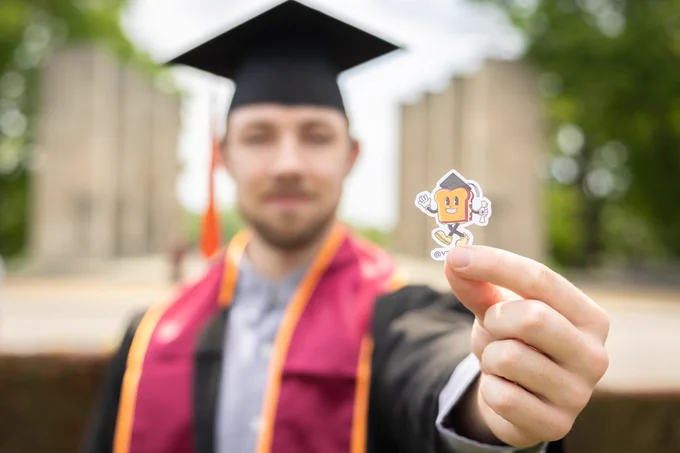 A Hokie in cap and gown stands in front of Pylons holding an exclusive graduate Cheesy Nights sticker. His tassel dangles from his hat and he has maroon and orange stole over his shoulders. There's green foliage in the background. 