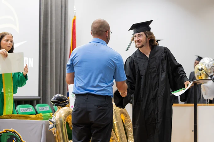 Graduate in black cap and gown shakes hands with a man in a blue shirt during a school ceremony on stage.
