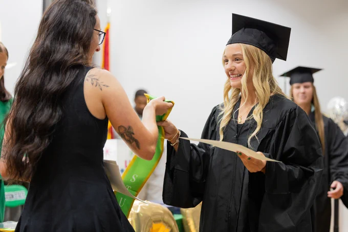 Graduate in cap and gown receiving diploma from a woman during a ceremony indoors.