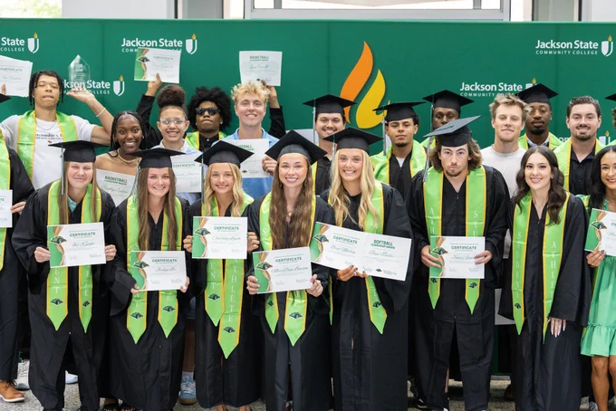 Group of graduates in black caps and gowns holding certificates in front of a Jackson State Community College backdrop.