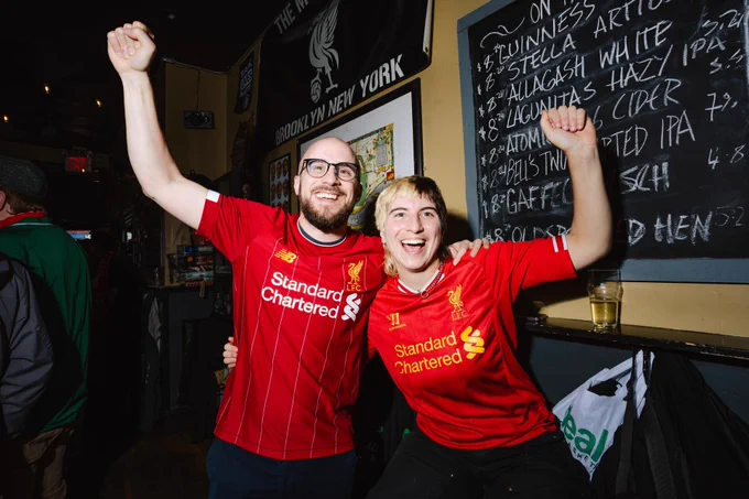 Two fans wearing Liverpool football jerseys cheer enthusiastically inside a pub with a beer menu on the wall behind them.