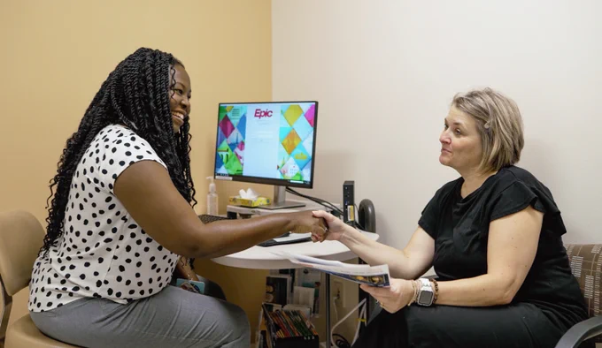 Monique R. Pappadis sits on the left in a clinic office and shakes hands with a seated woman holding printed materials. A computer monitor and office supplies are visible behind them.