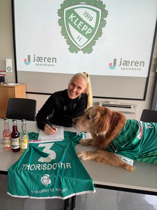 Maria Thorisdottir sat with a pen, paper, her dog Theo (in a Klepp shirt), and a shirt on the table with her surname and number 3 on, after signing for Klepp.
