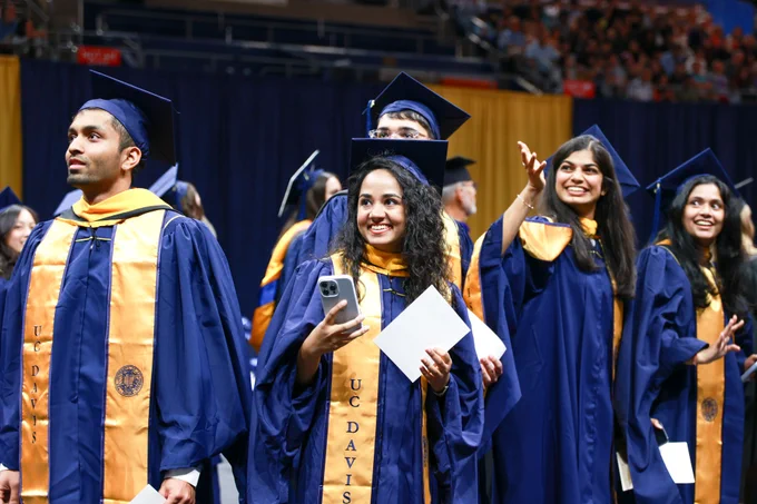 Graduate at Commencement smiling and holding up phone while others are looking around and smiling.