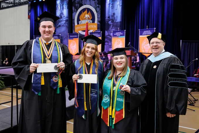 Three students in graduation attire are presented with awards from a university official. 