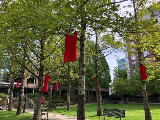 Red dresses hanging from tree branches in a green urban park with benches and buildings in the background.