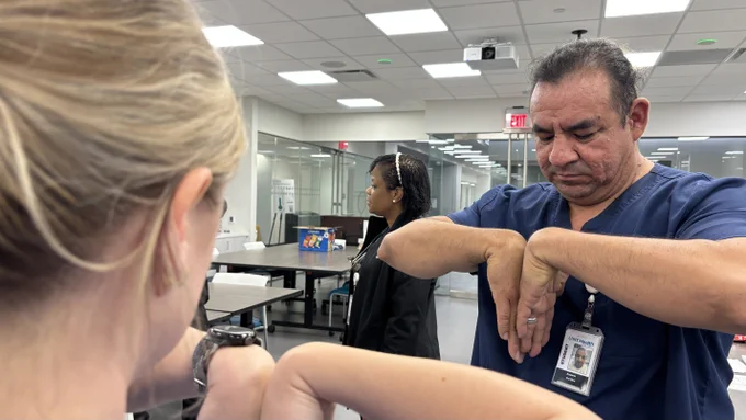 A person in medical attire demonstrates wrist exercises in a well-lit room with glass walls. Another individual watches attentively nearby, while a third person stands in the background.