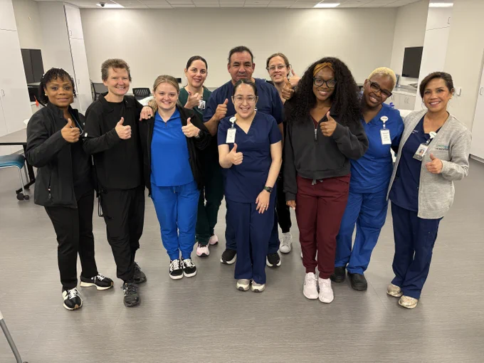A group of healthcare professionals in scrubs and uniforms are standing in a room, smiling, and giving thumbs up gestures. The environment appears to be a medical or clinical setting.