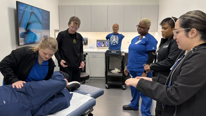 A group of healthcare professionals is in a medical training room. One person is examining a mannequin on a table, while others observe attentively. Medical posters and a torso model with visible organs are in the background. The room has a modern clinical appearance.