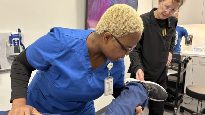 Healthcare professionals in a clinical setting assisting a patient with a leg examination. One person is wearing blue scrubs and is closely examining the patient's leg, while another in black attire provides additional support. Medical equipment is visible in the background.