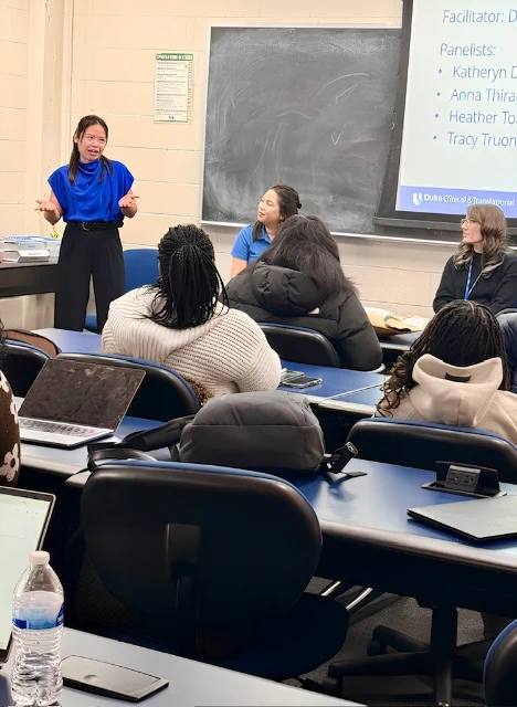 A classroom setting with a person standing and speaking, while two others sit and listen at the front of the room. The desks have laptops and a water bottle is visible. A presentation slide contains text with panelist names and includes the logo for Duke Clinical and Translational Science Institute.