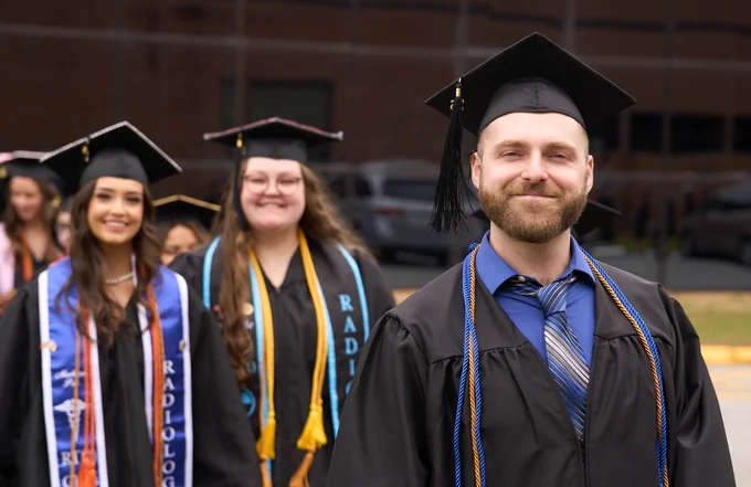 Smiling male graduate in cap and gown with two females behind walking in a line before ChattState graduation. 
