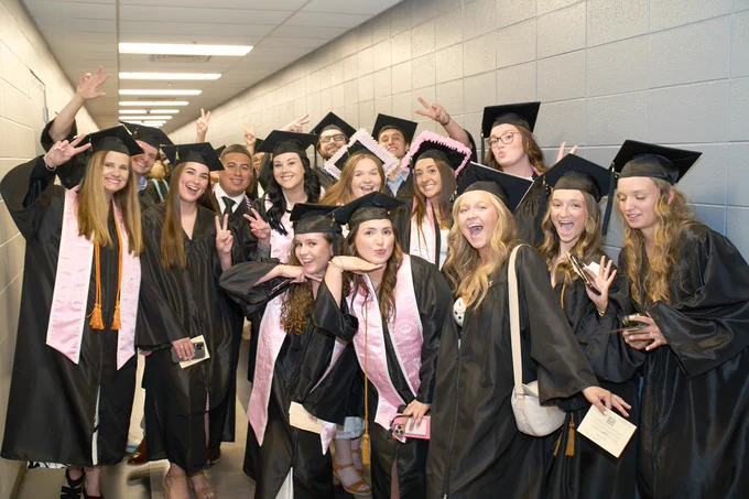 Group of RN graduates in black caps and gowns posing happily in a hallway, some wearing pink stoles, making peace signs and smiling at the camera.