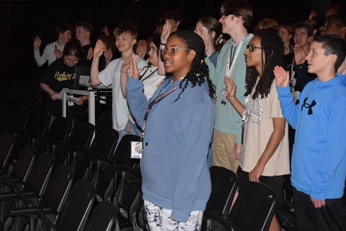 A group of students in an auditorium raise their right hands while reciting the pledge. Rows of black chairs fill the room, and a student in a blue hoodie stands prominently in the foreground.