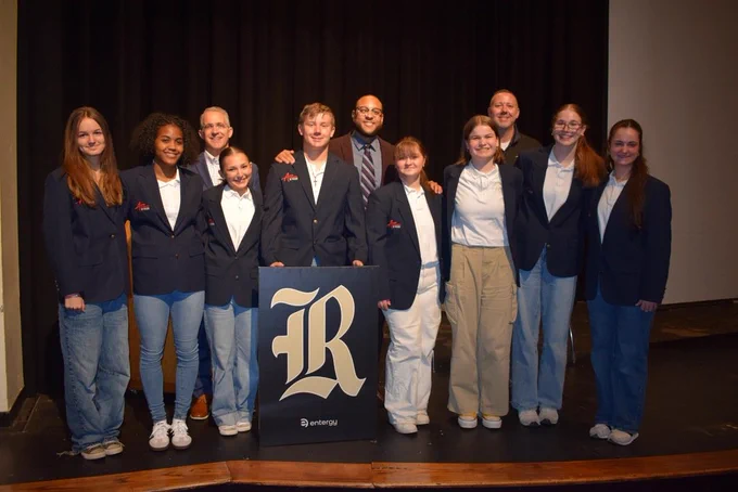 Ten individuals—eight students in matching navy blazers and two adults in suits—stand on a stage behind a podium featuring a large “R” and Entergy logo during the ceremony.