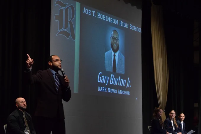 A speaker stands on stage with a microphone in front of a projected slide reading “Joe T. Robinson High School” and featuring a photo of Gary Burton Jr., labeled “KARK News Anchor.” Four seated panelists watch from the stage.