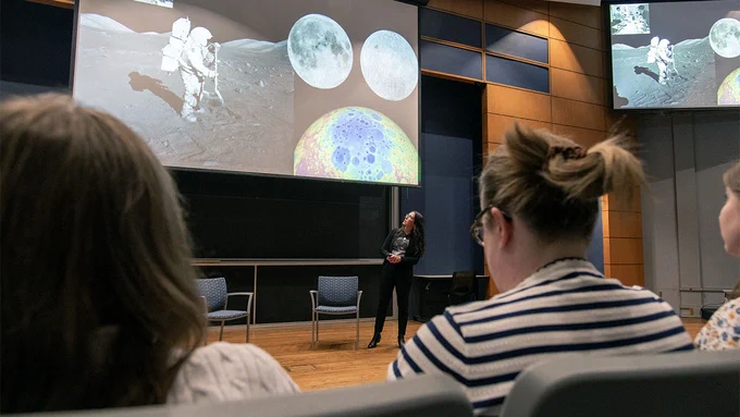 Kelsey Young gestures toward a screen displaying images of the Moon while audience members watch from their seats.