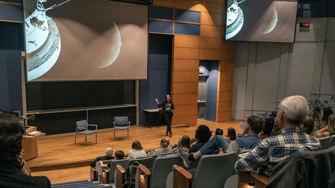Kelsey Young presents in a large lecture hall, with slides showing views of the Moon as attendees watch.