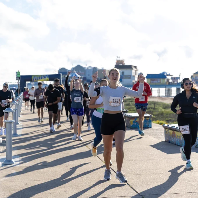 A group of people running a marathon on a paved path near a body of water. The sky is partly cloudy, and the runners are in athletic attire, with bib numbers pinned to their shirts.