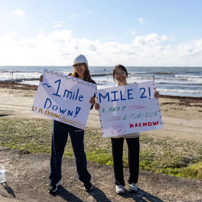 Two people are standing on a coastal path, each holding handmade signs. The water and beach are visible in the background under a clear blue sky.