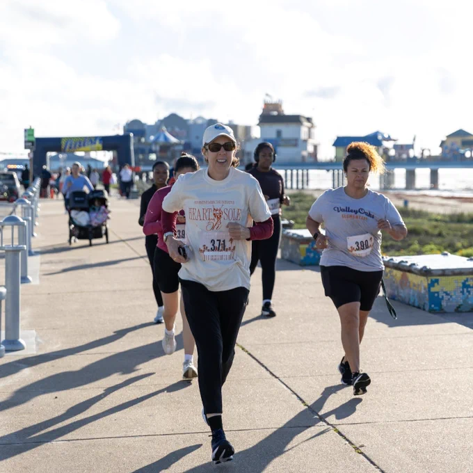 
People running in a charity race on a sunny day by the beach, with a Ferris wheel and other amusement park rides visible in the background.