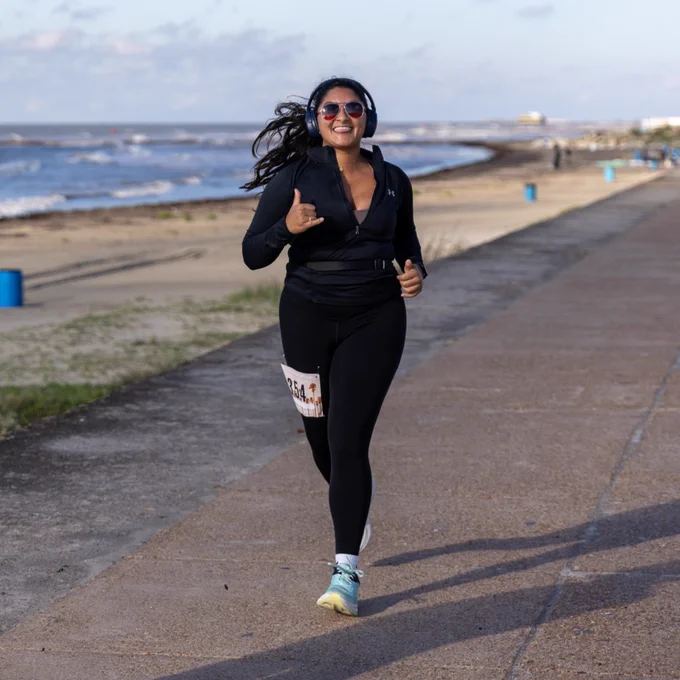 A person jogging along a beachside path, wearing headphones and athletic wear. The ocean is to the left, with a clear blue sky above.