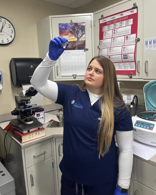 A person in a lab room holds up a small object, presumably for examination. They are wearing a navy uniform with a logo, blue gloves, and stand near laboratory equipment including a microscope and centrifuge. The wall features a scenic calendar image and safety notices.