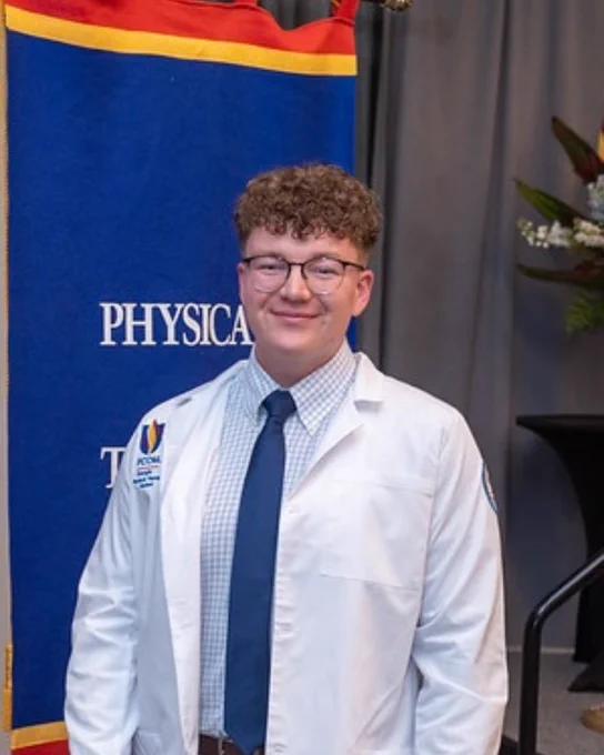 A person wearing a white coat and tie stands in front of a banner with the words "Physical." The background includes a table with a floral arrangement.