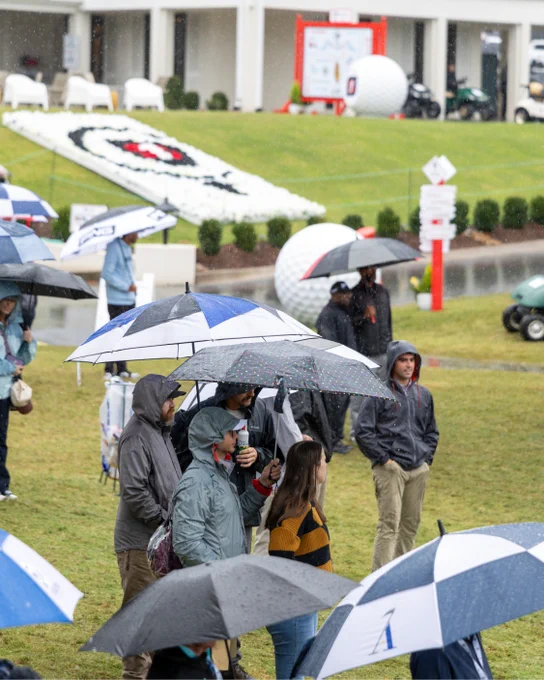 A group of people standing on grass, holding umbrellas on a rainy day. In the background, the Simmons Bank Championship logo is visible on a hillside.